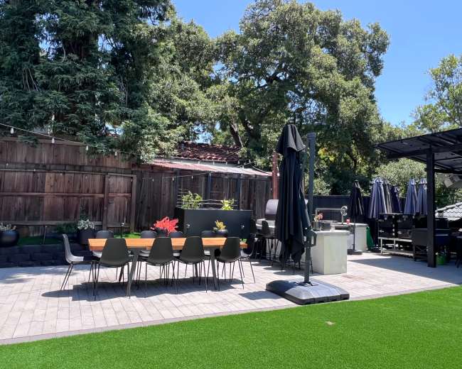 The image shows a landscaped outdoor patio with a long dining table, chairs, and several large umbrellas for shade, surrounded by greenery and trees.