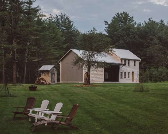 A wooden house with a metal roof sits amid a grassy yard surrounded by trees, featuring a small shed and four Adirondack chairs in the foreground.