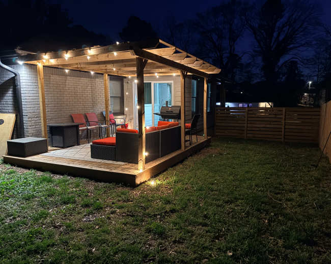 The image shows a well-lit outdoor patio with string lights, furnished with seating, and surrounded by grass and a wooden fence at night.