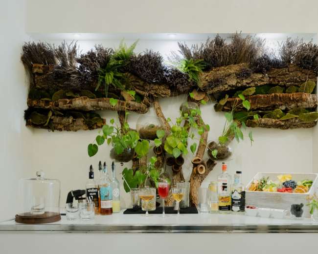 A modern bar setup features various bottles of liquor, glasses, and a display of fruits against a backdrop of decorative greenery and wooden elements.