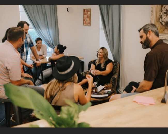 A group of people is engaged in conversation in a well-lit room with blue curtains and various seating arrangements.