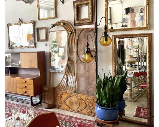 A vintage interior with a wooden furniture piece, multiple ornate mirrors, a decorative wall panel, plants in blue pots, and hanging light fixtures.