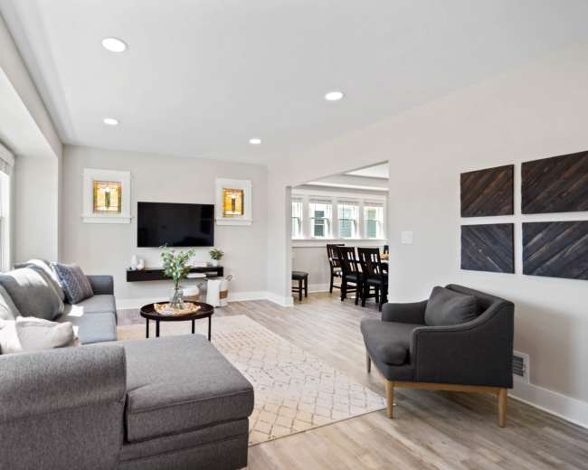 A modern living room features a gray sectional sofa, a round coffee table, and a TV mounted on the wall, with clear windows allowing natural light.