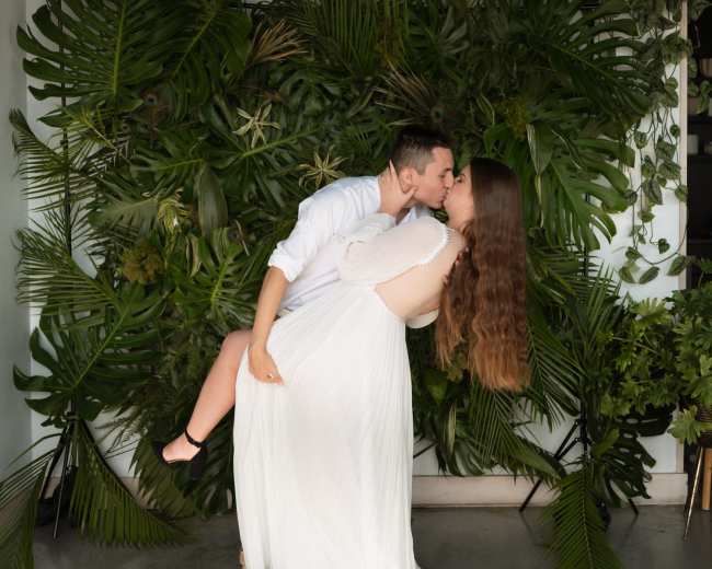 A couple shares a kiss while surrounded by lush green foliage in a modern indoor setting.