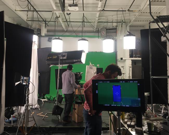 A film studio setup with a green screen background, several lights, and crew members preparing equipment.