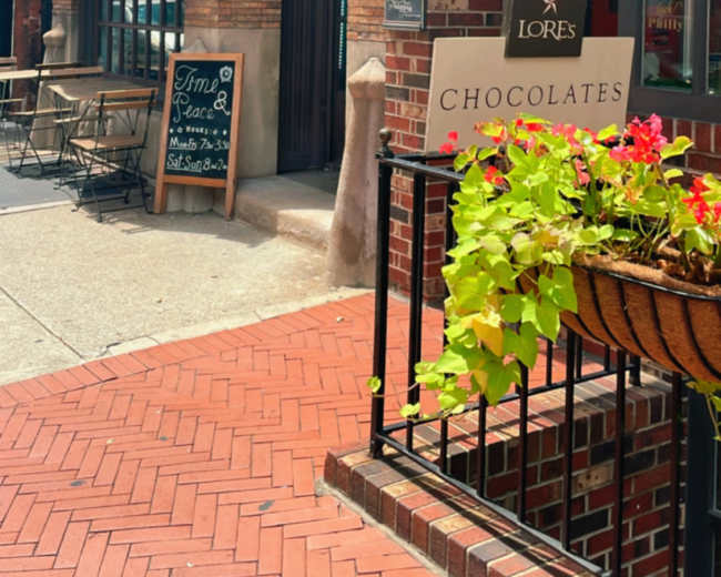 A brick storefront features a sign for "Lores Chocolates" beside a café with an open sign, while flower baskets adorn the entrance.