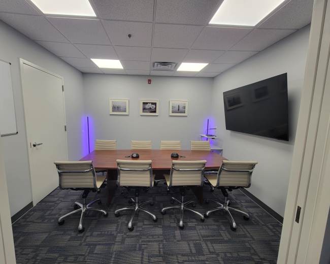 A modern conference room features a large wooden table surrounded by eight ergonomic chairs, a wall-mounted TV, and artwork, with lighting accents in purple.