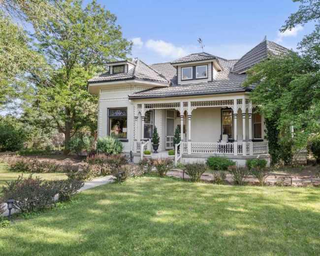 The image shows a large, two-story Victorian-style house surrounded by greenery and gardens.
