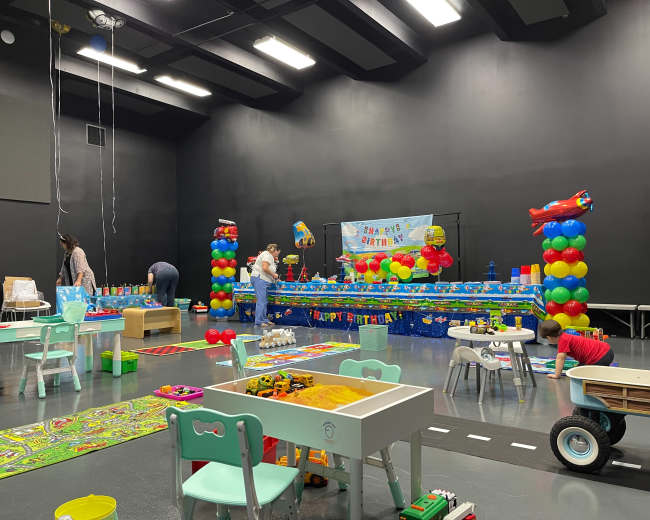 A children's party space is decorated with colorful balloons and a table filled with toys, while adults supervise various activities.