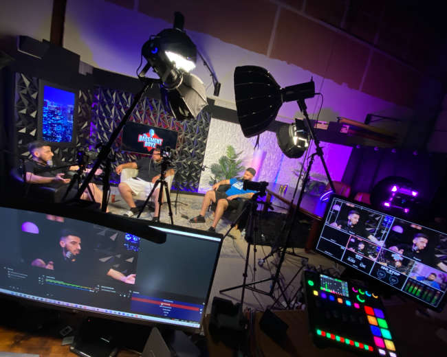 A recording studio setup with three individuals sitting on chairs, surrounded by cameras, lights, and a production console displaying live feeds.