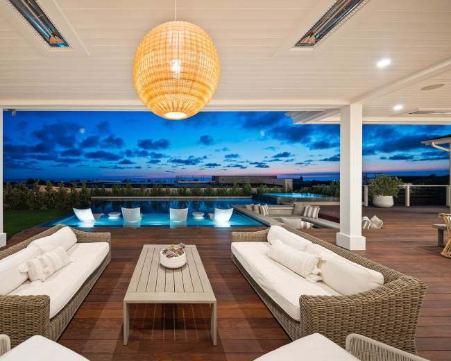 The image shows a modern outdoor living space with wicker furniture arranged around a wooden coffee table, overlooking a pool under a twilight sky.