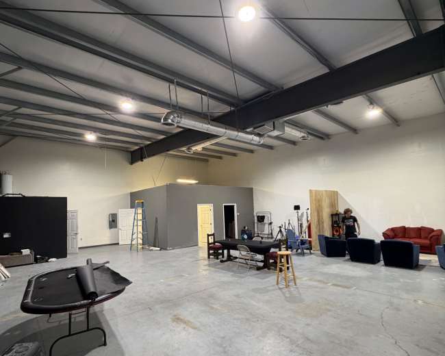 The image shows an empty industrial-style studio space with exposed beams, concrete flooring, and various furniture and equipment scattered around.