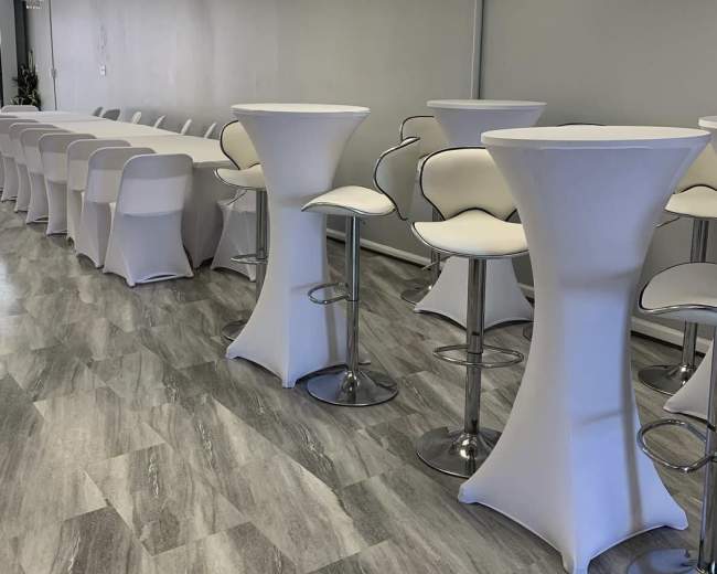 A spacious event room featuring white cocktail tables and high-backed stools arranged neatly along a light-colored wall.