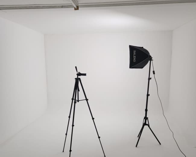 The image shows a minimal photography studio setup featuring a tripod and a softbox light in a white room.