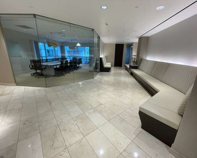 An office lobby features tiled flooring, sleek seating along the walls, and a glass-walled meeting room in the background.