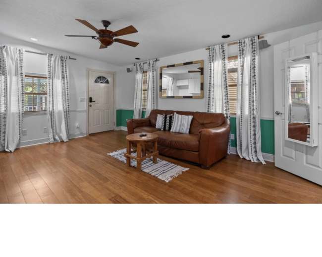 The image shows a living room with a brown leather sofa, a small wooden table, and large windows dressed with striped curtains.