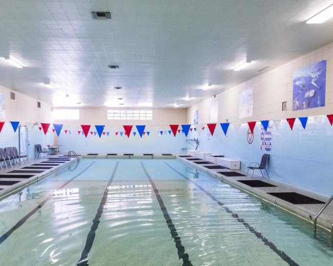 An indoor swimming pool with red, white, and blue triangular flags hung above the water and seating arranged along the side.