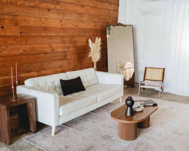 The image shows a modern living room with a white leather sofa, a wooden accent wall, a round coffee table, and a large mirror reflecting the space.