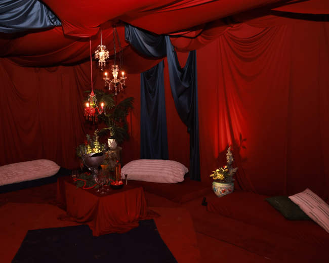 The image shows a dimly lit, red-themed tent interior with draped fabric, a low table adorned with decorative items, and cushioned seating on the floor.