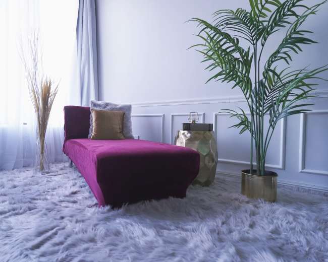 The image shows a purple chaise lounge placed on a plush, white area rug, next to a potted plant and a small side table with glassware.