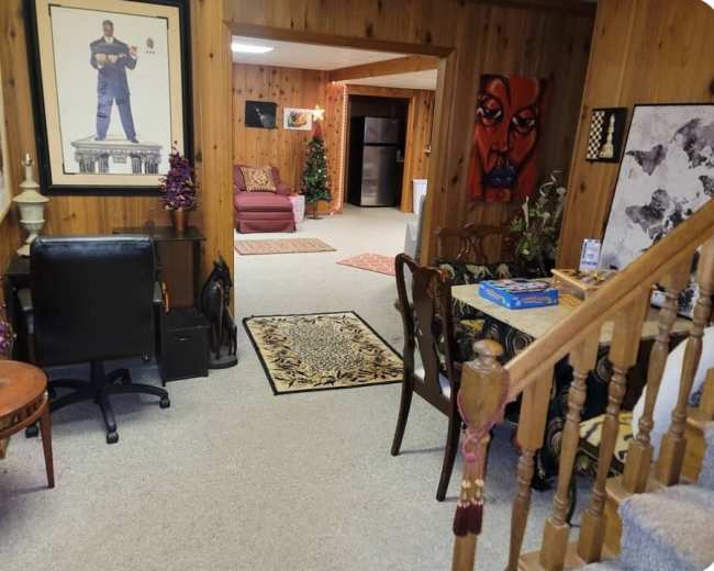 The image shows a cozy basement area featuring wooden paneling, a seating area with a couch, a small table with a game, and various decorations, including artwork and a Christmas tree.