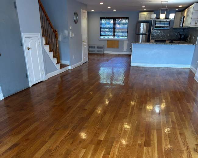The image shows a spacious, empty living area with polished hardwood floors and a kitchen area in the background.