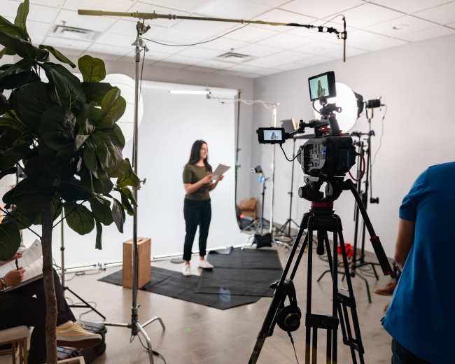 A woman is standing on a black mat in front of a white backdrop while holding a script, surrounded by video equipment and studio lights.