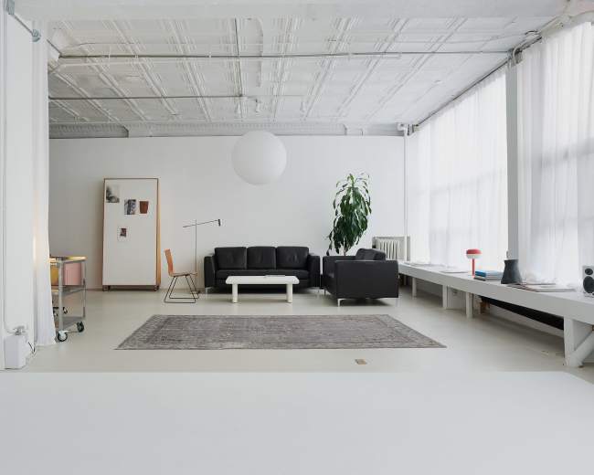 The image shows a spacious, minimalist living area with a black sofa, a white coffee table, and large windows allowing natural light to filter through.