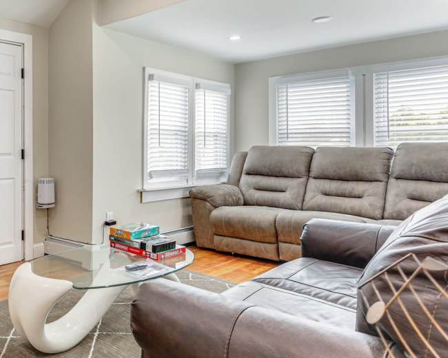 The image shows a modern living room with a gray sectional sofa, a glass coffee table, and large windows with white blinds.