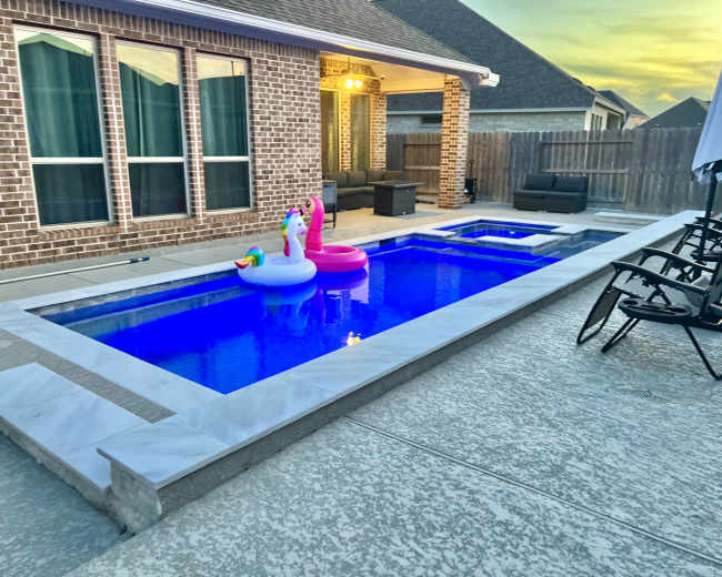 A modern backyard features a rectangular pool with colorful inflatable floats, surrounded by a patio area with chairs and a seating arrangement.
