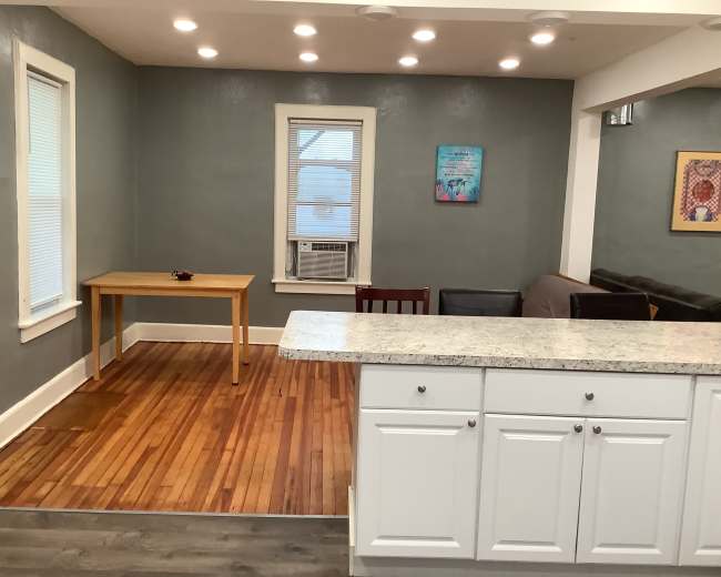 A kitchen area with a white countertop and cabinets, adjacent to a small dining table and chairs, set against a wall with light-colored paint and wood flooring.