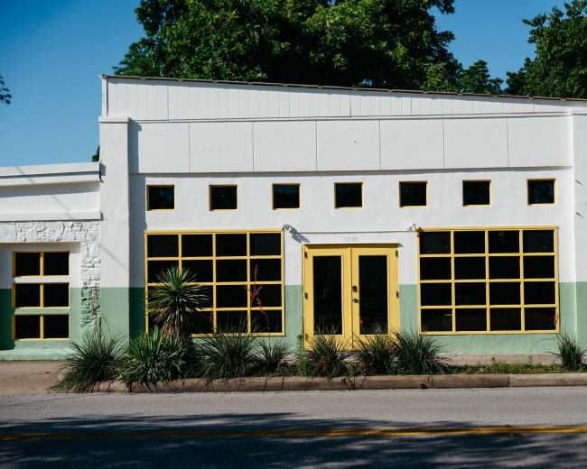 The building features a white exterior with yellow trim and large windows on the front facade, set against a backdrop of greenery.