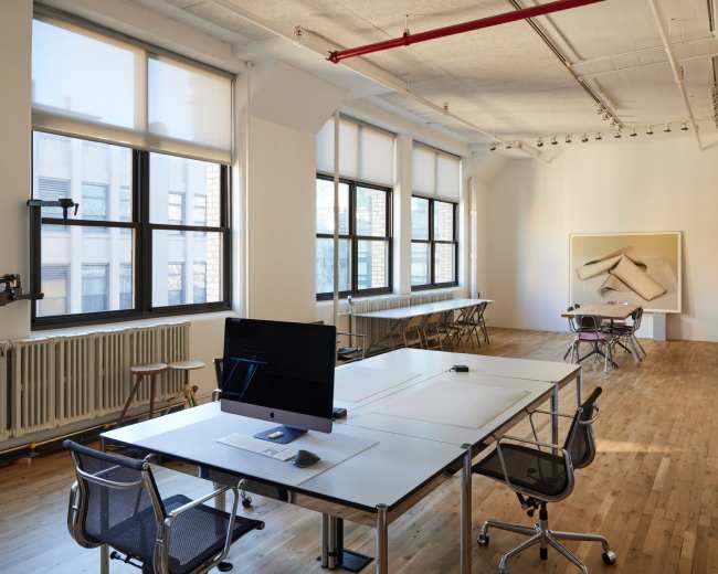 The image shows a bright, spacious office with large windows, wooden floors, and modern furniture including a desk with a computer.