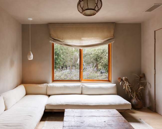 A minimalist living room features a L-shaped white couch, a wooden coffee table, and a large window overlooking greenery.