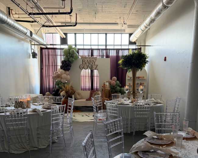 The image shows a decorated event space featuring several tables with clear chairs, a backdrop with the words "Baby Gomez," and various decorative elements, including balloons and plants.