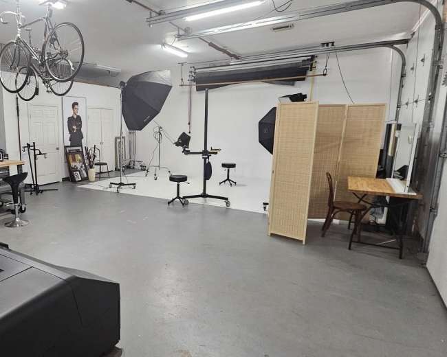The image shows a well-lit photography studio with backdrop equipment, light stands, and a workspace featuring chairs and a table.