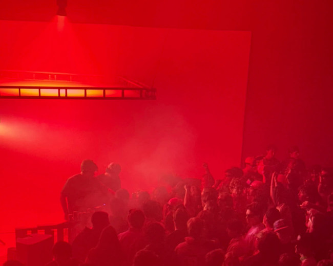 A dense crowd gathers under a red-lit atmosphere with fog as a DJ performs on stage.