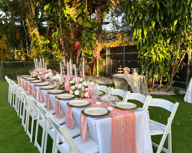 A long banquet table is elegantly set up outdoors, adorned with pink table runners, floral centerpieces, and candles, surrounded by lush greenery.