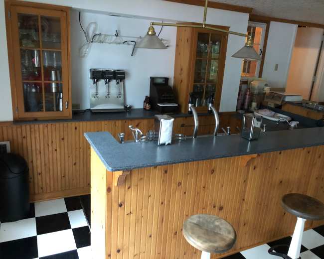 The image shows a wooden bar area with a countertop, three stools, and cabinets, set against a black and white checkered floor.