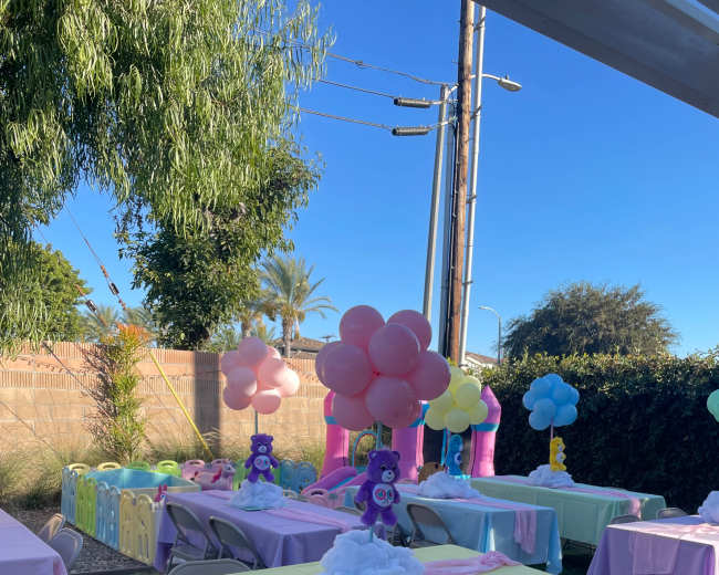 The image shows a colorful outdoor party setup with tables covered in pastel linens and decorative balloons in various colors.