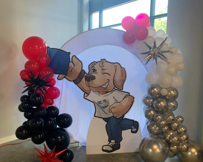 The scene features a balloon arch with black, red, and silver balloons surrounding a cutout of a cartoon dog character holding a blue object.