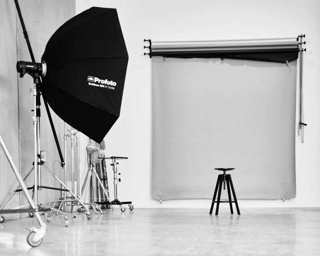 The image shows a photography studio with a black umbrella light, a gray backdrop, and a black stool in the center.