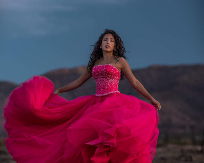 A woman in a vibrant pink gown twirls with a mountainous landscape in the background.