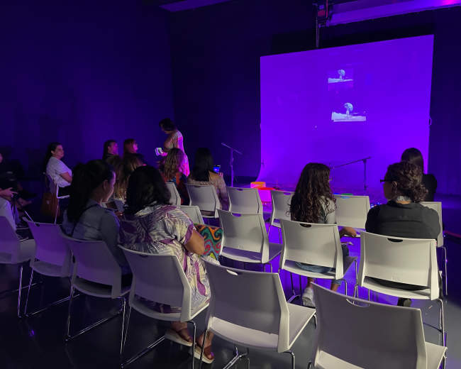 A group of people sits in white chairs facing a large screen displaying an image, with a stage and microphone in the background.