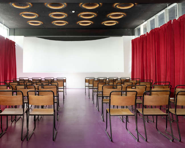 The image shows a spacious seminar room with rows of metal-framed wooden chairs facing a blank white screen, flanked by red curtains.