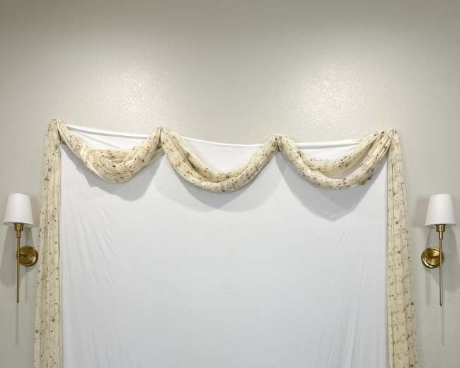 The image shows a plain white backdrop draped with a floral-patterned fabric, flanked by two wall sconces, in front of a wooden stool.
