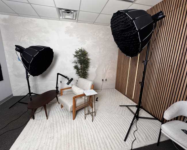 The image shows a studio setup with two large softbox lights, a wooden table, an armchair, and a plant against a textured wall.