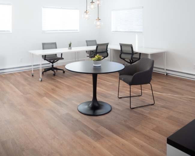 The image shows a modern office space with two tables, four chairs, and hanging pendant lights, featuring a wooden floor and white walls.