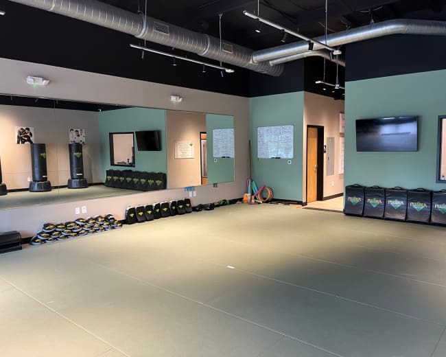The image shows a fitness studio with a padded floor, mirrored walls, wall-mounted television, and various workout equipment neatly arranged along the sides.