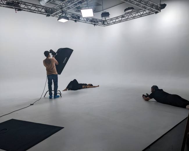 A photographer uses a softbox while a model lies on the ground in a white studio with two assistants positioned around them.